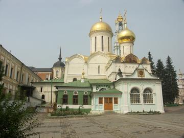 Белая церковь с золотыми главами посреди монастырского двора, рядом низкие постройки с зелёными крышами и длинный жёлто-белый корпус