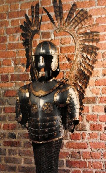 На фоне кирпичной стены стоит стальной доспех с нагрудником из полос и шлемом, за спиной поднимаются полукругом два ряда тёмных перьев на высоких древках