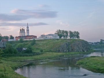 На скалистом берегу реки стоят белые каменные постройки кремля с собором и колокольней, впереди река и зелёный луг