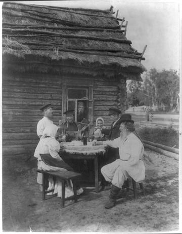 Старая чёрно-белая фотография: у бревенчатой избы с соломенной крышей за столом с самоваром сидят и стоят крестьяне — седобородый старик, женщины в платках, молодые парни в картузах, один играет на балалайке