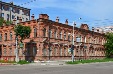 Двухэтажное здание из красного кирпича на углу улицы, окна с решётками, у входа небольшая табличка