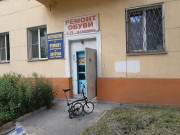 Жилой дом с вывеской «Ремонт обуви» над открытой дверью мастерской, у входа стоит велосипед